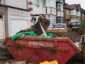 A residential street scene with a row of semi-detached houses. In the foreground, a large, red skip bin filled with construction debris, including wood, dirt, a green tarp, and patterned fabric, is situated on a driveway. Two cars are parked further back in front of the houses, which have a mix of decorative facades and bay windows.