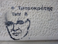 A stencil graffiti on a textured wall features a simple, black outline of a person's face alongside Greek text. The wall has a rough, beige surface with visible textures and shadows.