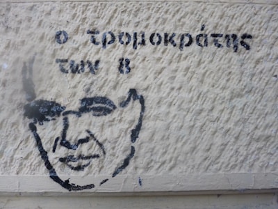 A stencil graffiti on a textured wall features a simple, black outline of a person's face alongside Greek text. The wall has a rough, beige surface with visible textures and shadows.