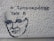 A stencil graffiti on a textured wall features a simple, black outline of a person's face alongside Greek text. The wall has a rough, beige surface with visible textures and shadows.