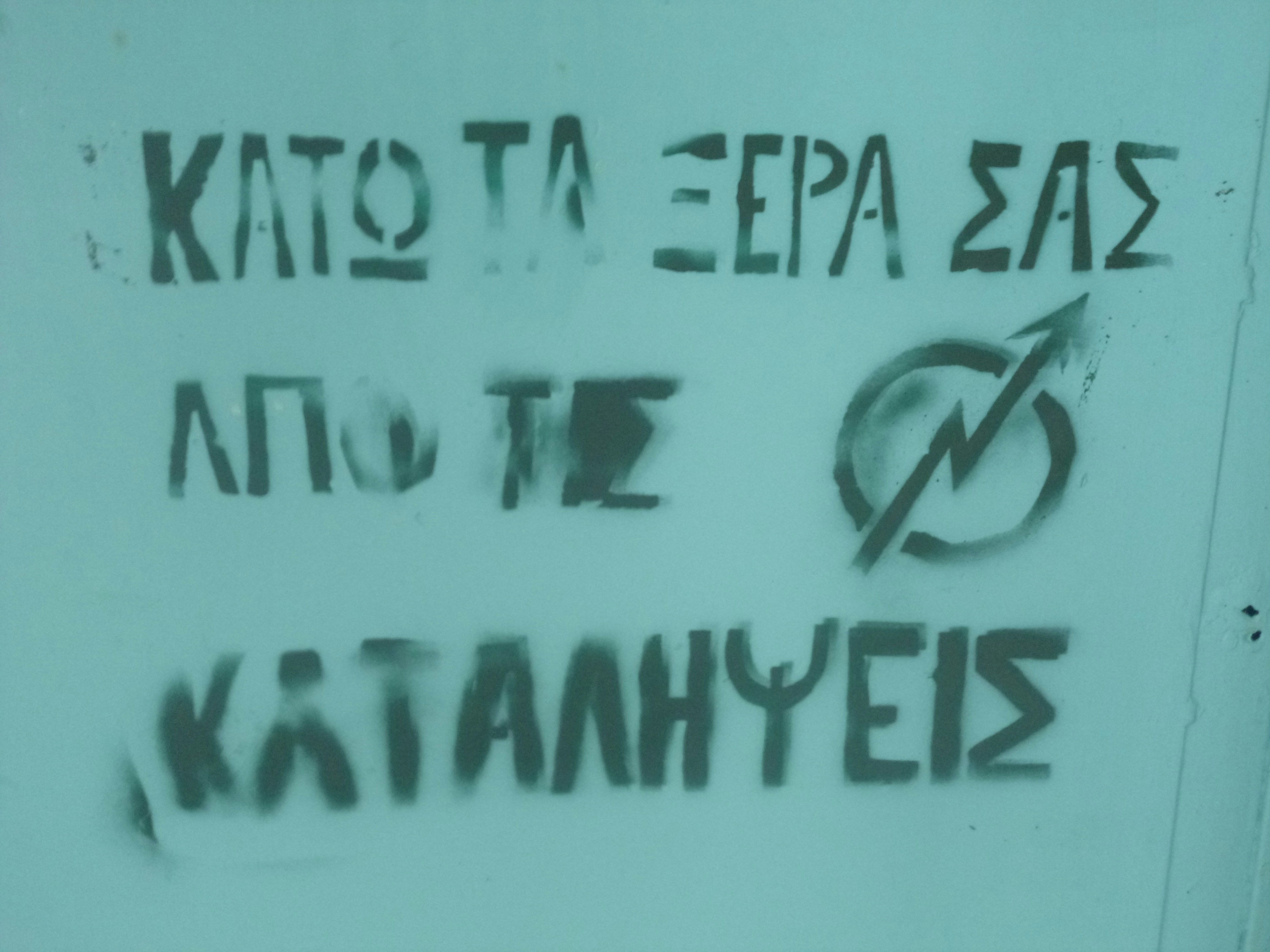 Graffiti on a light-colored wall with Greek text and a symbol resembling an arrow inside a circle with a slash. The text appears to be a political or protest statement.