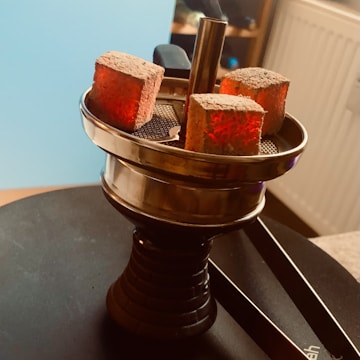 A hookah top with three glowing charcoal cubes set on a metal tray, creating a warm and ambient atmosphere. The background shows a radiator and a part of a room setting.