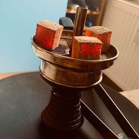 A hookah top with three glowing charcoal cubes set on a metal tray, creating a warm and ambient atmosphere. The background shows a radiator and a part of a room setting.