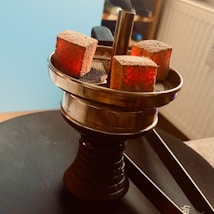 A hookah top with three glowing charcoal cubes set on a metal tray, creating a warm and ambient atmosphere. The background shows a radiator and a part of a room setting.