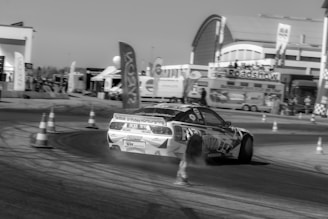 A sports car is drifting around a track surrounded by traffic cones. The background features a building with signs and banners, indicating an event or roadshow. The scene appears to be at an organized racing or motorsport event.