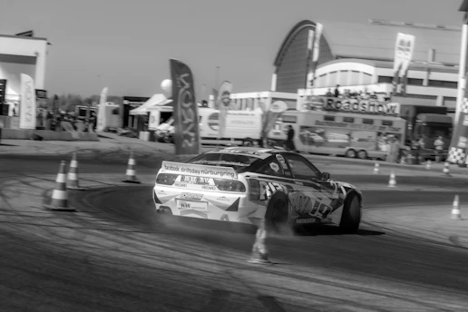 A sports car is drifting around a track surrounded by traffic cones. The background features a building with signs and banners, indicating an event or roadshow. The scene appears to be at an organized racing or motorsport event.
