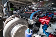 A close-up view of a high-performance car engine is visible, showcasing intricate components like a large turbocharger, colorful belts and pulleys, and various metal fittings. The setting appears to be a mechanic's garage or workshop, with metal beams and lighting fixtures above, and blurred background elements suggesting other machinery or vehicles.