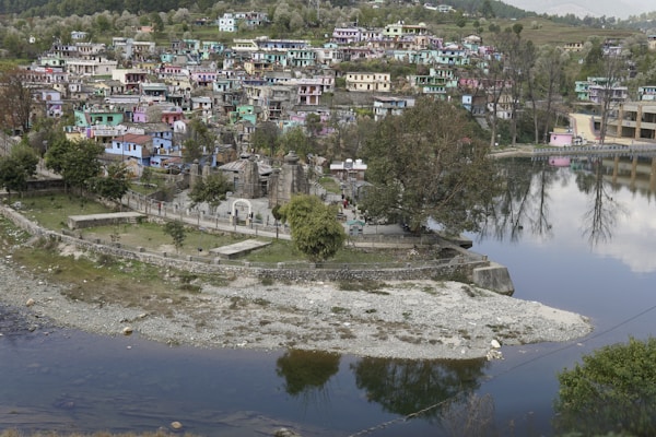 A picturesque town with colorful houses nestled on a hillside, reflected in a calm body of water. The area is lush with trees, and a cluster of ancient-looking stone structures or temples is situated near the water's edge. The town appears vibrant and lively, with a mix of modern and traditional architecture.