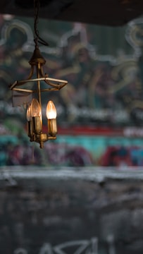 A vintage-style chandelier with several bulbs hanging against a blurred, graffiti-covered wall. The chandelier has a rustic, brass appearance with exposed bulbs providing a warm glow.