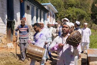 A group of people dressed in traditional attire participating in a festive procession outside a rural building. Some individuals are playing musical instruments like drums, and others have colorful powder on their clothes and faces. The bright sunlight enhances the vividness of the colors.