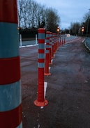 A row of red and white striped traffic bollards lines the side of a road, with trees in the background and street lights illuminating the scene. The road appears to be slightly wet, suggesting recent rain, and the sky is overcast, adding a gloomy atmosphere.