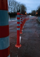 A row of red and white striped traffic bollards lines the side of a road, with trees in the background and street lights illuminating the scene. The road appears to be slightly wet, suggesting recent rain, and the sky is overcast, adding a gloomy atmosphere.