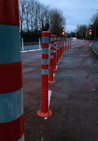 A row of red and white striped traffic bollards lines the side of a road, with trees in the background and street lights illuminating the scene. The road appears to be slightly wet, suggesting recent rain, and the sky is overcast, adding a gloomy atmosphere.