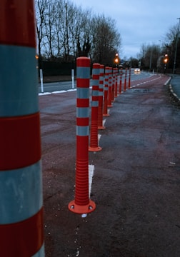 A row of red and white striped traffic bollards lines the side of a road, with trees in the background and street lights illuminating the scene. The road appears to be slightly wet, suggesting recent rain, and the sky is overcast, adding a gloomy atmosphere.