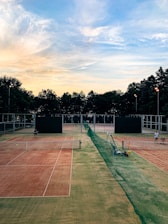 A serene tennis court setting during sunset. Two clay courts are visible, divided by a green net fence. There are two players preparing for a match, each standing on a different court. The sky is filled with soft pastel clouds transitioning from light blue to a warm yellow-orange near the horizon. Trees line the background, silhouetted against the colorful sky.