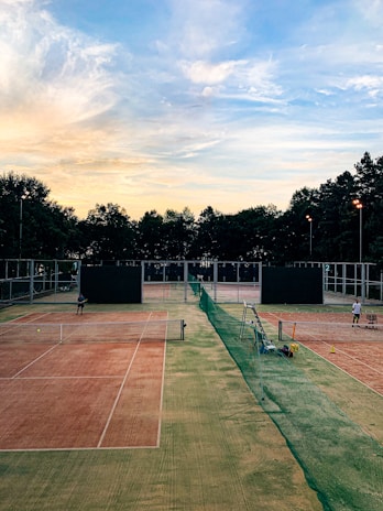 A serene tennis court setting during sunset. Two clay courts are visible, divided by a green net fence. There are two players preparing for a match, each standing on a different court. The sky is filled with soft pastel clouds transitioning from light blue to a warm yellow-orange near the horizon. Trees line the background, silhouetted against the colorful sky.
