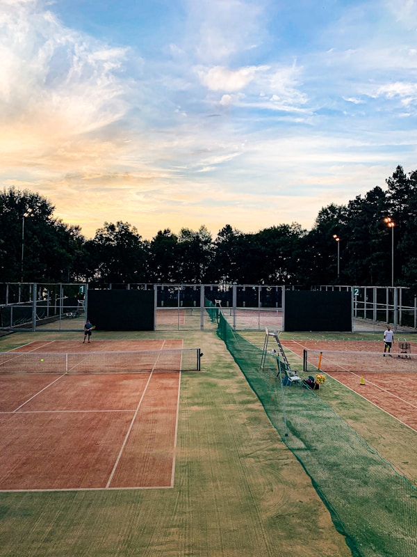 A serene tennis court setting during sunset. Two clay courts are visible, divided by a green net fence. There are two players preparing for a match, each standing on a different court. The sky is filled with soft pastel clouds transitioning from light blue to a warm yellow-orange near the horizon. Trees line the background, silhouetted against the colorful sky.