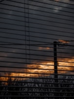 Horizontal window blinds partially obscure a view of an outdoor scene with a cloudy sky and a sunset or sunrise, casting a warm orange and yellow glow through the slats. A silhouette of a pole or tree is visible against the diminishing light.