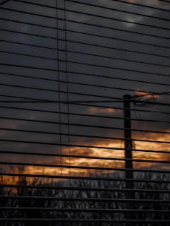 Horizontal window blinds partially obscure a view of an outdoor scene with a cloudy sky and a sunset or sunrise, casting a warm orange and yellow glow through the slats. A silhouette of a pole or tree is visible against the diminishing light.