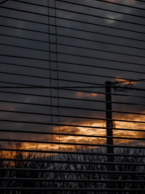 Horizontal window blinds partially obscure a view of an outdoor scene with a cloudy sky and a sunset or sunrise, casting a warm orange and yellow glow through the slats. A silhouette of a pole or tree is visible against the diminishing light.