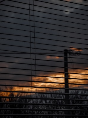 Horizontal window blinds partially obscure a view of an outdoor scene with a cloudy sky and a sunset or sunrise, casting a warm orange and yellow glow through the slats. A silhouette of a pole or tree is visible against the diminishing light.