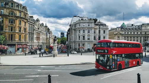 A busy urban scene featuring iconic London architecture, with a prominent red double-decker bus turning onto a city street. Historic buildings line the streets on both sides, and a statue stands in a square area. People can be seen walking along the sidewalks, and traffic flows through the intersection. The sky is partly cloudy.