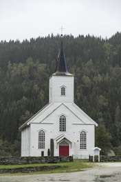 A white wooden church with a tall steeple topped with a cross, surrounded by a dense green forest. The church has red doors and several large, arched windows. The setting is serene, with overcast skies contributing to a calm atmosphere.