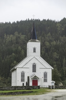 A white wooden church with a tall steeple topped with a cross, surrounded by a dense green forest. The church has red doors and several large, arched windows. The setting is serene, with overcast skies contributing to a calm atmosphere.