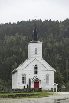 A white wooden church with a tall steeple topped with a cross, surrounded by a dense green forest. The church has red doors and several large, arched windows. The setting is serene, with overcast skies contributing to a calm atmosphere.