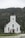 A white wooden church with a tall steeple topped with a cross, surrounded by a dense green forest. The church has red doors and several large, arched windows. The setting is serene, with overcast skies contributing to a calm atmosphere.