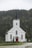 A white wooden church with a tall steeple topped with a cross, surrounded by a dense green forest. The church has red doors and several large, arched windows. The setting is serene, with overcast skies contributing to a calm atmosphere.