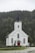 A white wooden church with a tall steeple topped with a cross, surrounded by a dense green forest. The church has red doors and several large, arched windows. The setting is serene, with overcast skies contributing to a calm atmosphere.