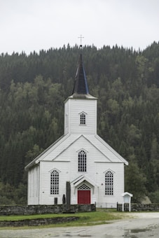 A white wooden church with a tall steeple topped with a cross, surrounded by a dense green forest. The church has red doors and several large, arched windows. The setting is serene, with overcast skies contributing to a calm atmosphere.