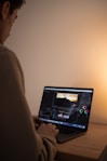 A person is seated at a wooden desk, focused on editing a video. The open laptop screen displays video editing software featuring various tools and timeline sections. The lighting is soft and warm, creating a relaxed ambiance.