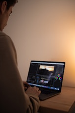 A person is seated at a wooden desk, focused on editing a video. The open laptop screen displays video editing software featuring various tools and timeline sections. The lighting is soft and warm, creating a relaxed ambiance.