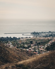 An expansive landscape featuring a suburban area nestled between rolling hills and the vast ocean. Rows of houses with red and beige roofs are visible alongside well-maintained green gardens. The coastline stretches out with breakwaters extending into the calm sea. The overall scene conveys a sense of tranquility and orderliness.