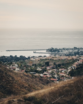 An expansive landscape featuring a suburban area nestled between rolling hills and the vast ocean. Rows of houses with red and beige roofs are visible alongside well-maintained green gardens. The coastline stretches out with breakwaters extending into the calm sea. The overall scene conveys a sense of tranquility and orderliness.