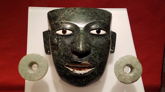 A polished, ancient-looking mask carved from dark green stone, with intricate eye details and a subtle expression. Flanking the mask are two circular jade ornaments. The background is a rich red color.