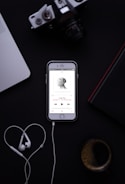 A smartphone displaying a music app with earphones placed below it, forming a heart shape. The background includes a laptop, a camera, a book, and a cup of coffee.