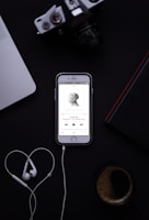 A smartphone displaying a music app with earphones placed below it, forming a heart shape. The background includes a laptop, a camera, a book, and a cup of coffee.
