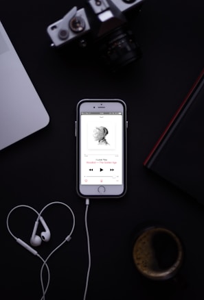 A smartphone displaying a music app with earphones placed below it, forming a heart shape. The background includes a laptop, a camera, a book, and a cup of coffee.