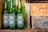 Three green glass Heineken beer bottles are displayed against a background of rustic bricks. The bottles are standing upright on a wooden or stone surface, with their labels prominently featuring the brand name and logo.