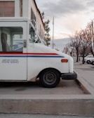 A parked United States Postal Service truck is positioned on a concrete pavement. The vehicle is white with a blue and red stripe along the side, and it is set against the background of a building and bare trees on a quiet street.