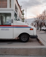 A parked United States Postal Service truck is positioned on a concrete pavement. The vehicle is white with a blue and red stripe along the side, and it is set against the background of a building and bare trees on a quiet street.