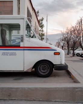 A parked United States Postal Service truck is positioned on a concrete pavement. The vehicle is white with a blue and red stripe along the side, and it is set against the background of a building and bare trees on a quiet street.