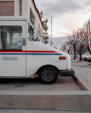 A parked United States Postal Service truck is positioned on a concrete pavement. The vehicle is white with a blue and red stripe along the side, and it is set against the background of a building and bare trees on a quiet street.