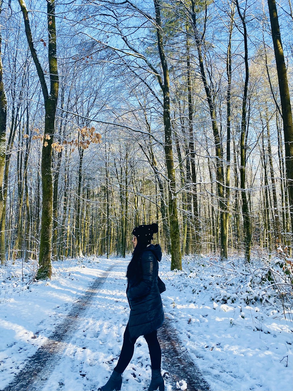 person in black jacket walking on snow covered pathway between bare trees during daytime