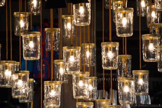A collection of vintage-style glass jars with glowing light bulbs inside, suspended from the ceiling with brown cords. The light cast by the bulbs creates a warm and inviting glow, with reflections caught on the glass surfaces. Surrounding elements are blurred, highlighting the artistic lighting arrangement.