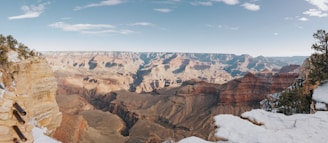 A vast, expansive view of the Grand Canyon, showcasing its deep ravines and numerous rock formations carved by the Colorado River. The scene is bathed in natural light, highlighting the stratified layers of rock in hues of red, orange, and brown. Snow-capped edges and surrounding greenery enhance the natural beauty and ruggedness of the landscape.