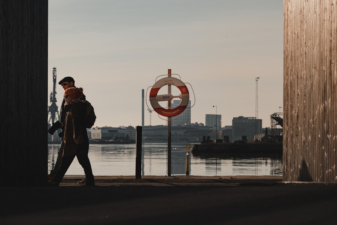 man in black jacket and black pants standing on sidewalk near body of water during daytime